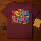 Maroon t-shirt featuring "Cousin Camp 2026" with cartoon sun, trees, tent, and campfire.